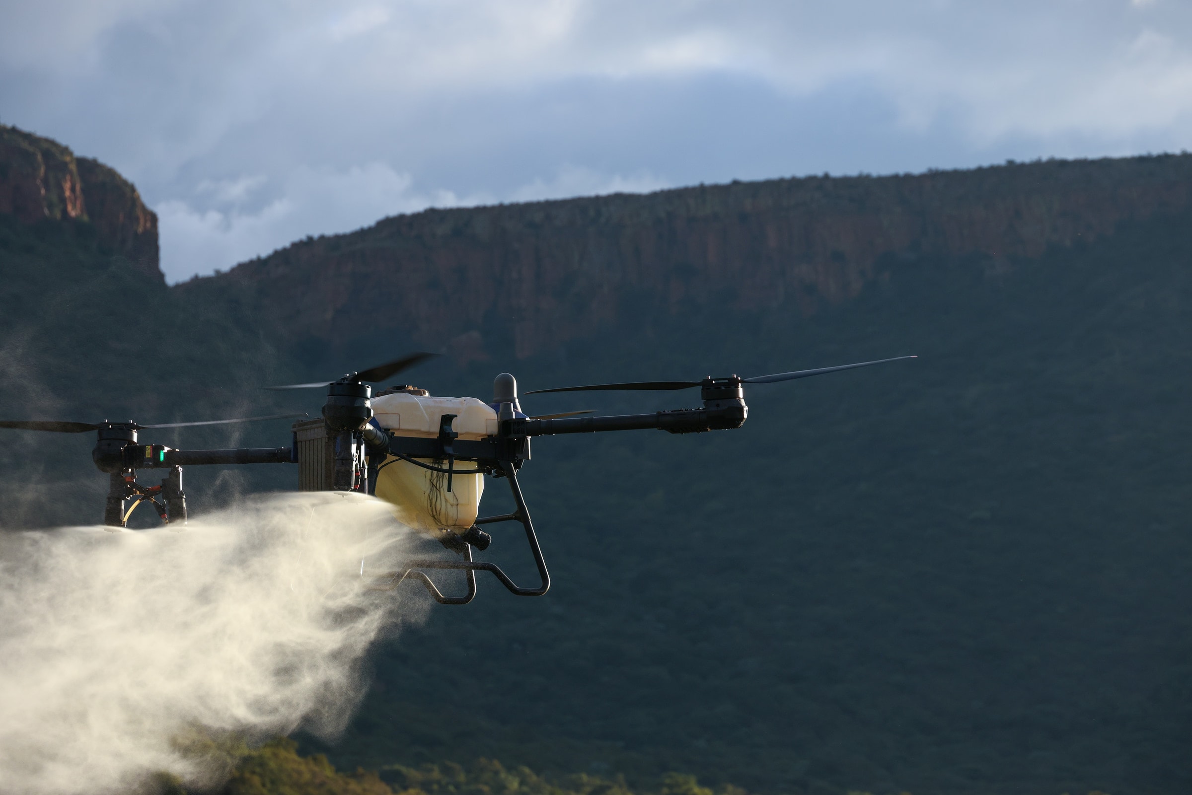 Drone spraying chemicals over a scenic mountainous landscape under a cloudy sky.