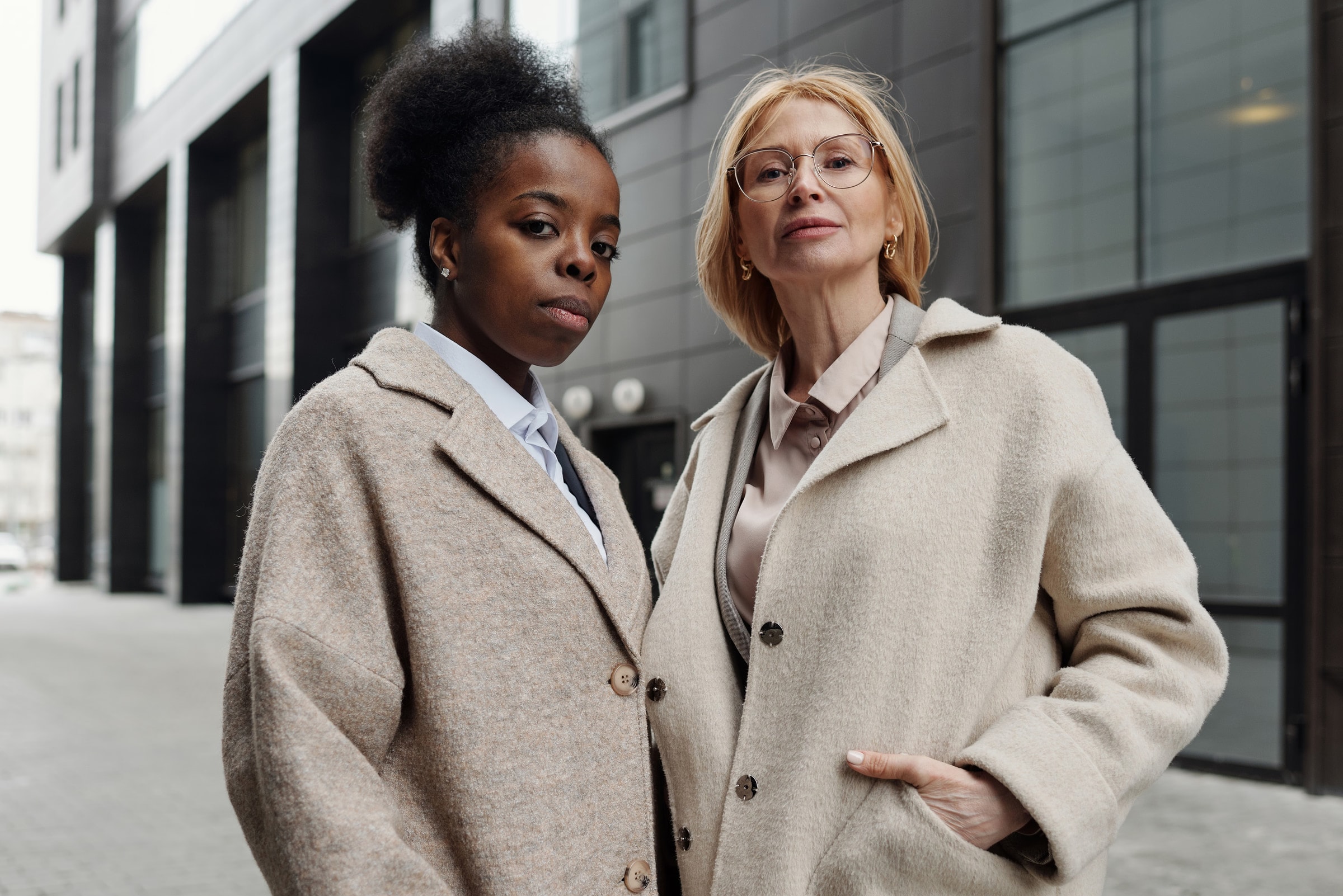 Two diverse businesswomen in stylish coats posing confidently on a city street.